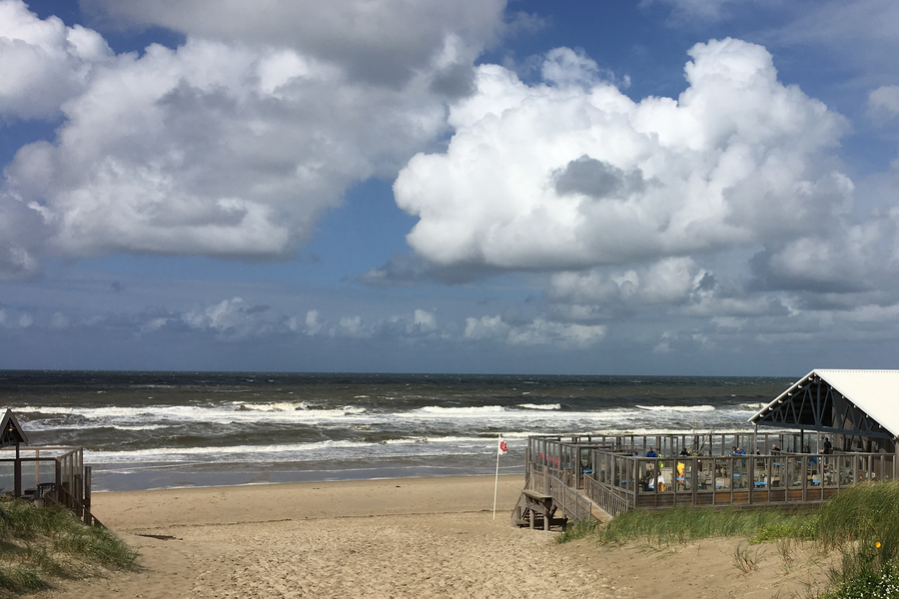 Foto mit Strand, Strandpavillion und Wellen der Nordsee, darüber blauer Himmel mit weißen Wolken